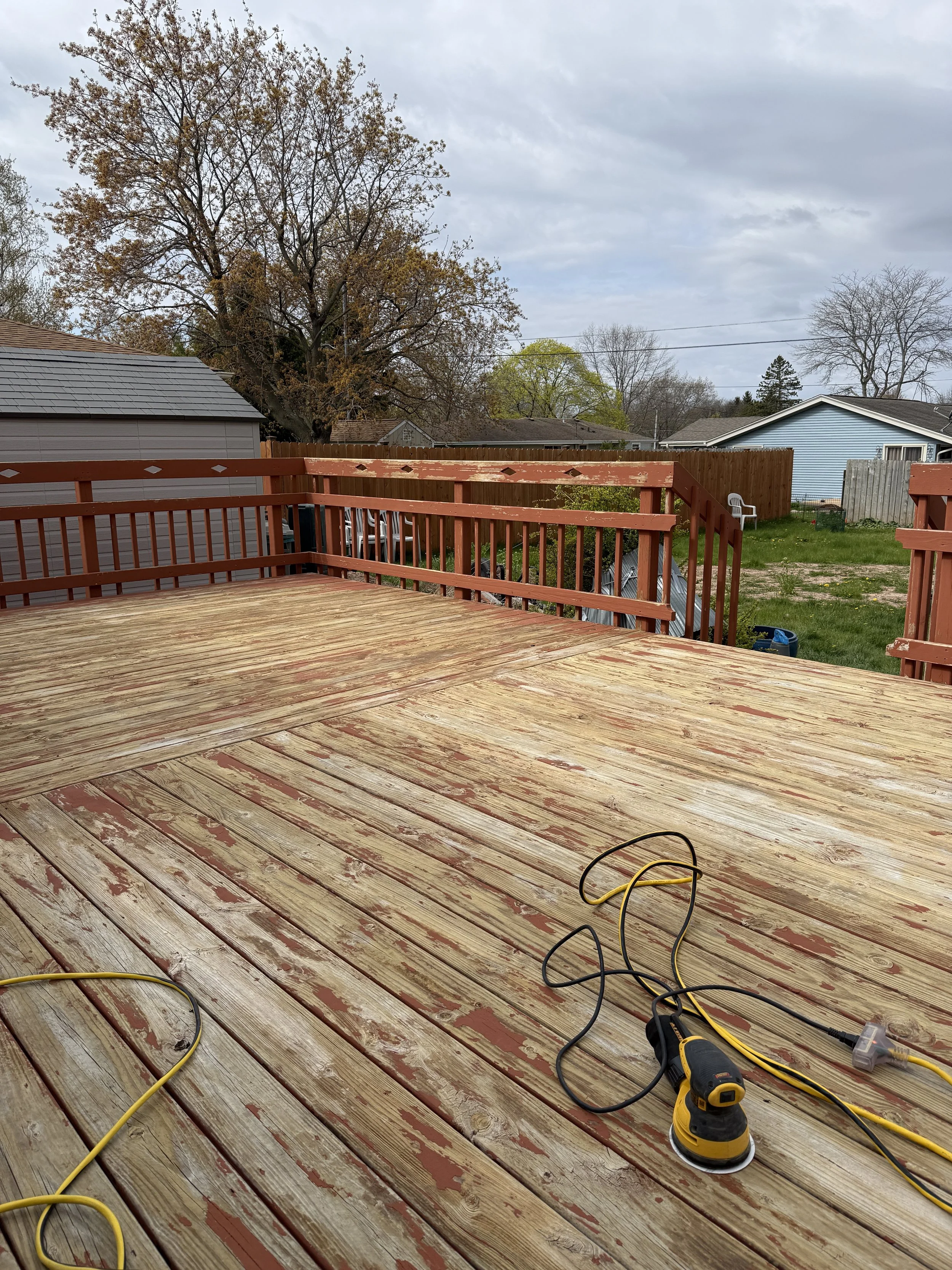 A wooden deck under construction with power tools and extension cords on the surface, with a backyard featuring trees, a lawn, and neighboring houses visible in the background under a cloudy sky.