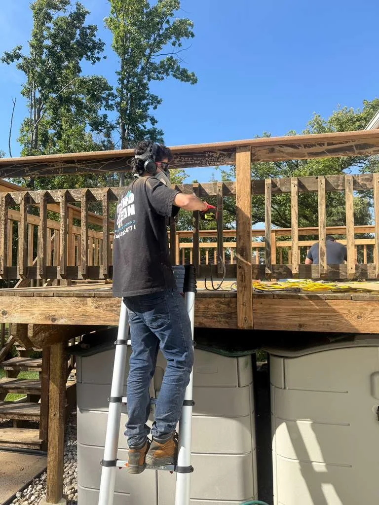 A person standing on a ladder working on a wooden deck, with another person in the background, trees and a clear blue sky.