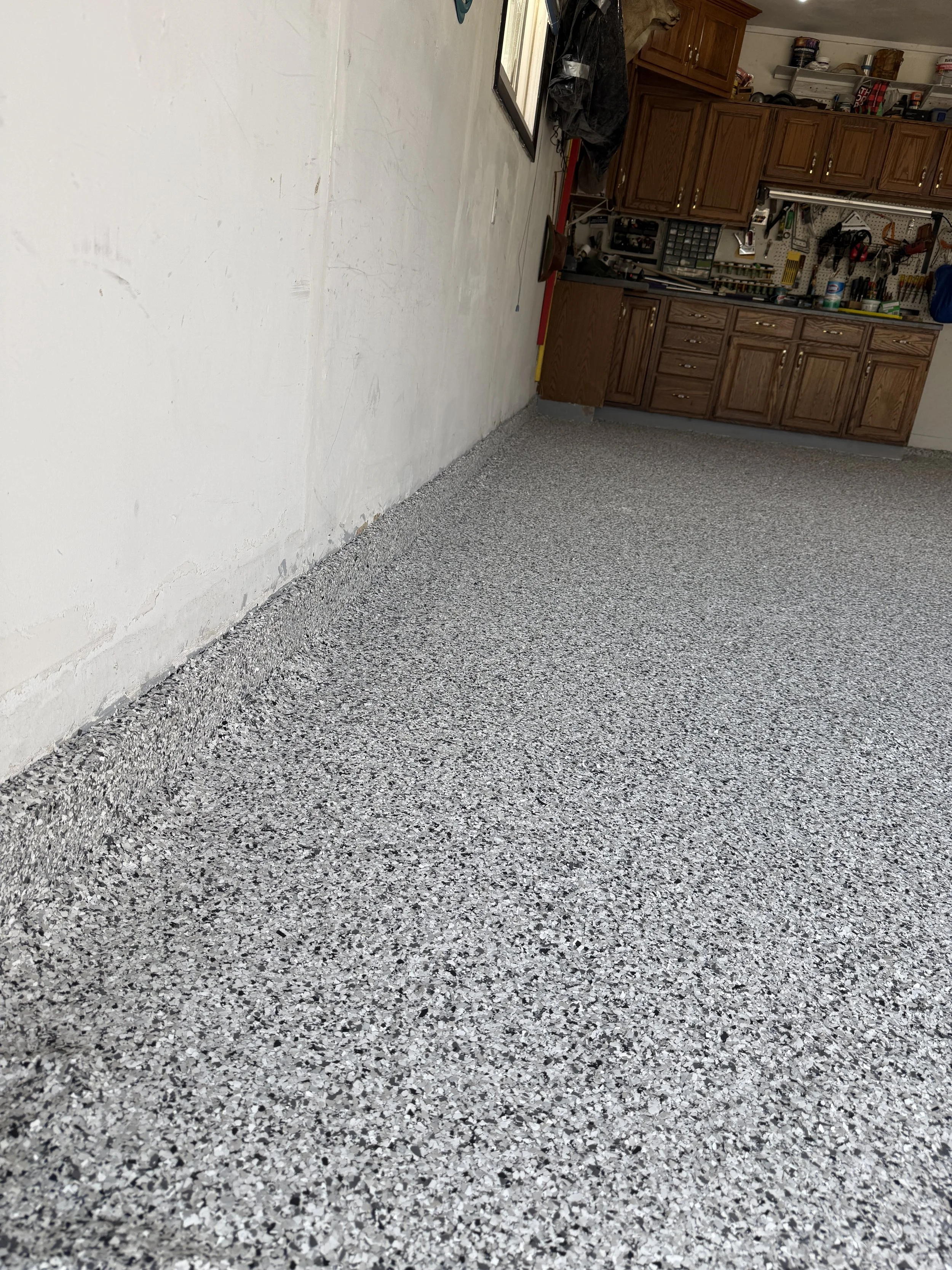 Photo of a garage or workshop with a speckled epoxy floor, a white brick wall, and wooden cabinets filled with tools and supplies.