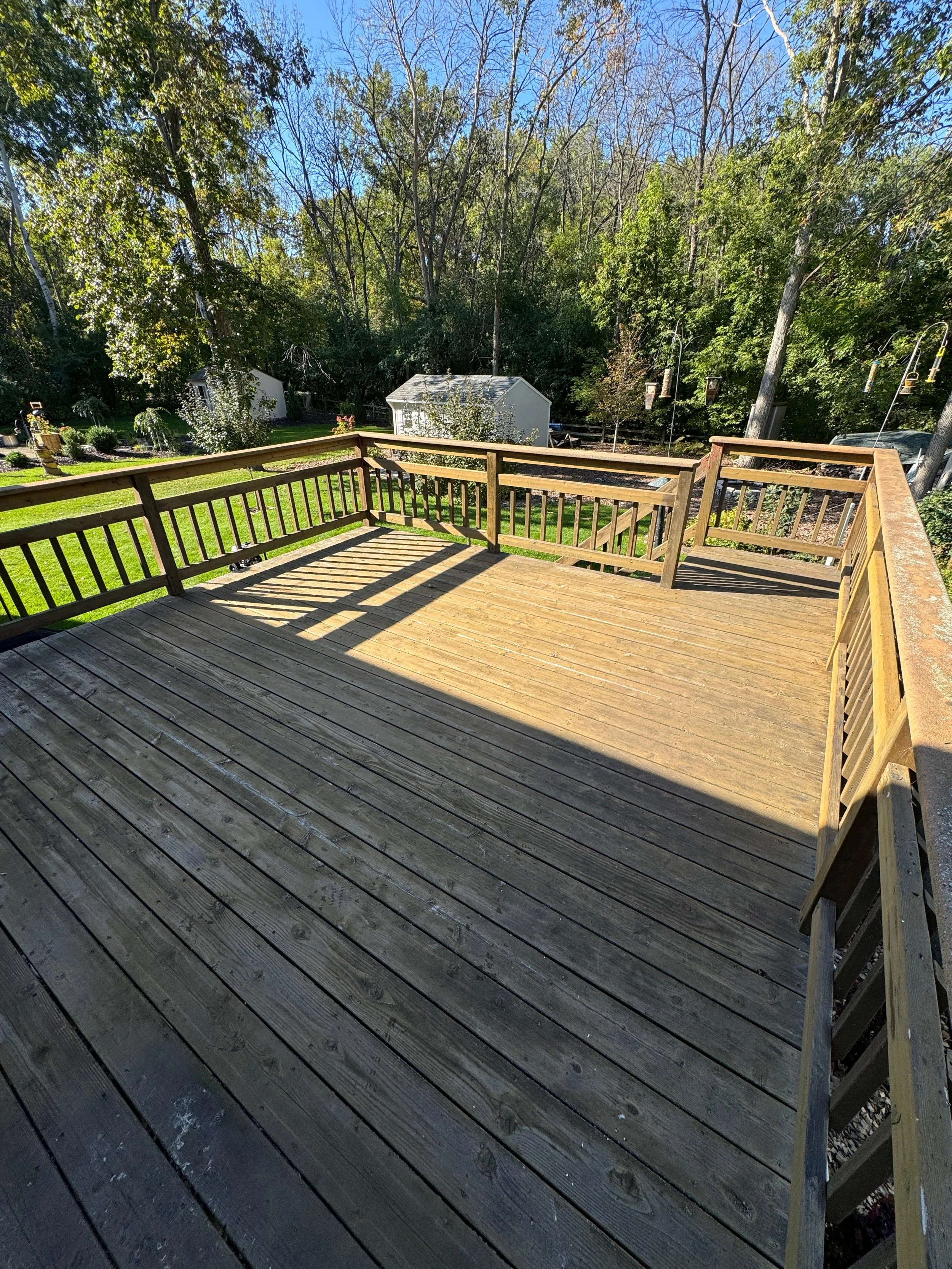 Wooden outdoor deck with railing, shadowed area, backyard with trees, small shed, and garden in the background.