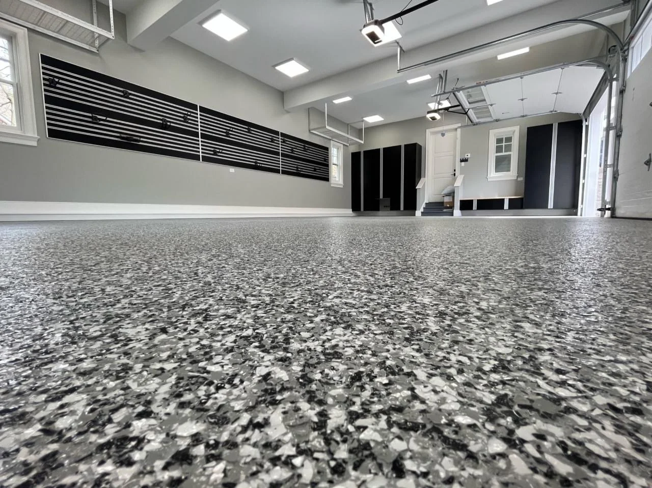 Empty garage with speckled gray and black epoxy floor, gray walls, black storage cabinets, and garage door open to outside.
