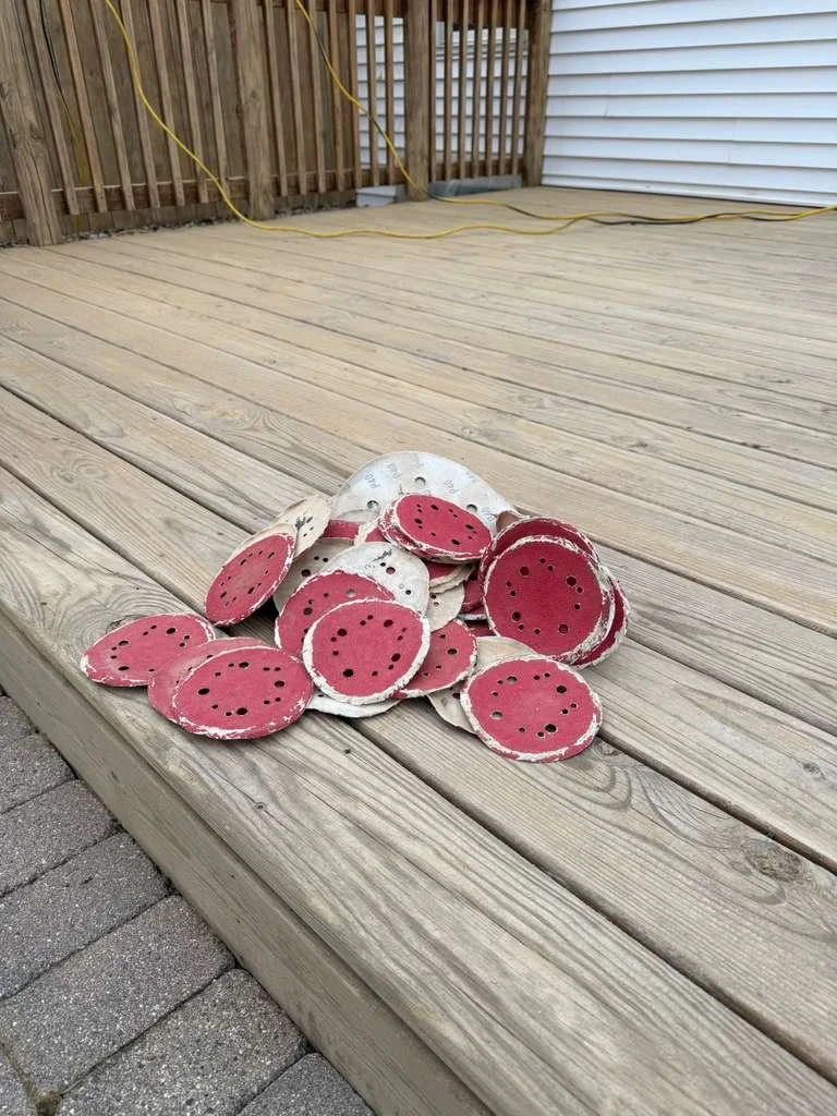 Pile of round ceramic watermelon slice decorations on a wooden deck, with a background of a wooden fence and a white building.
