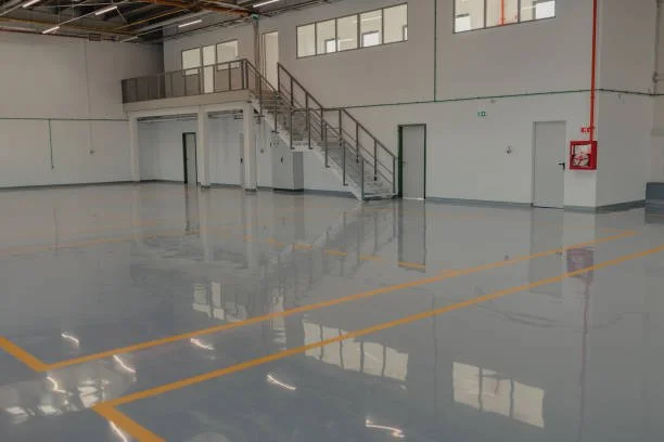 Empty industrial or warehouse space with polished concrete floor, metal staircase leading to a mezzanine, enclosed offices or rooms with windows, and safety equipment on wall.