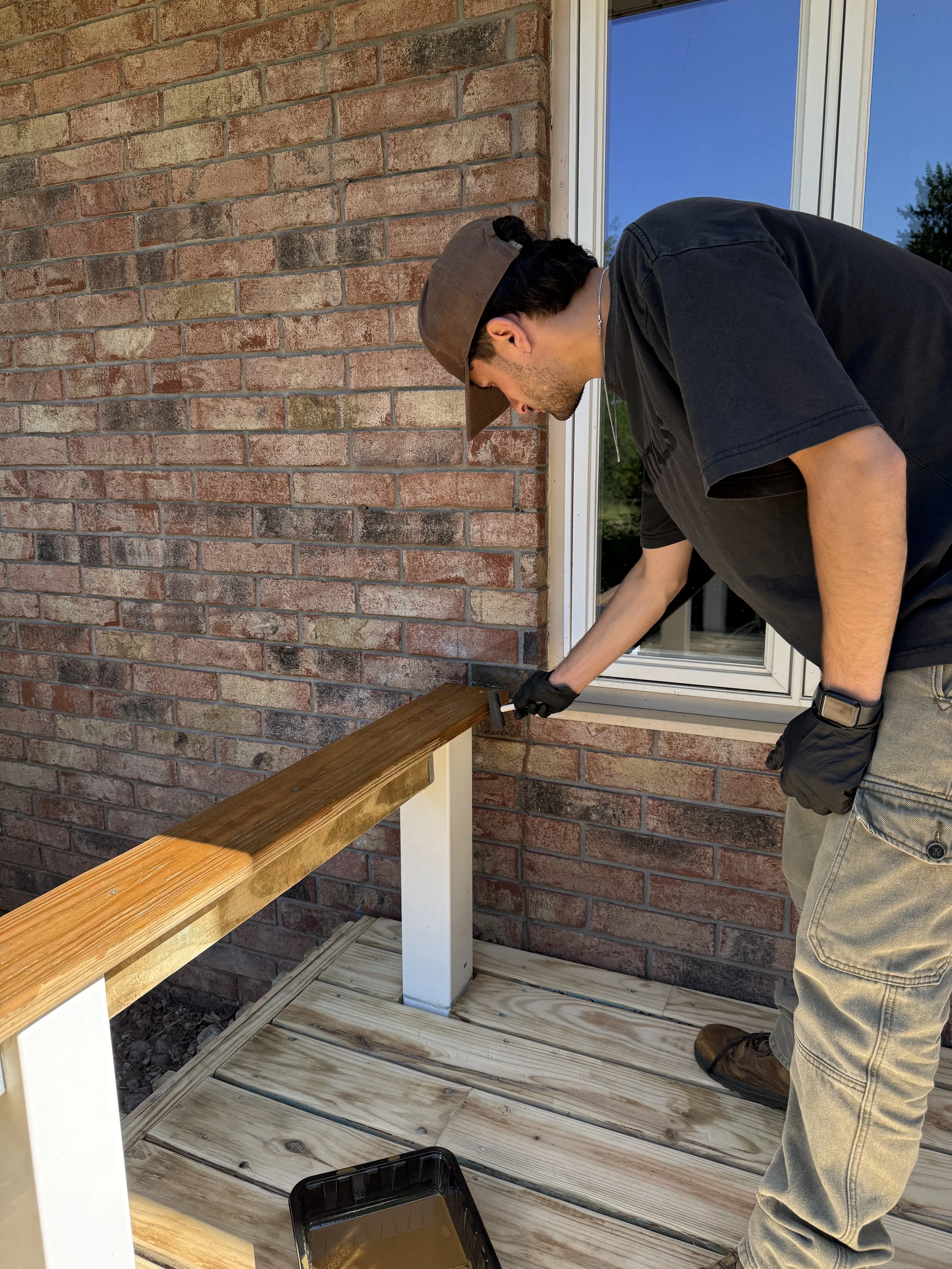 A man staining a wooden railing on a porch next to a brick wall and window.