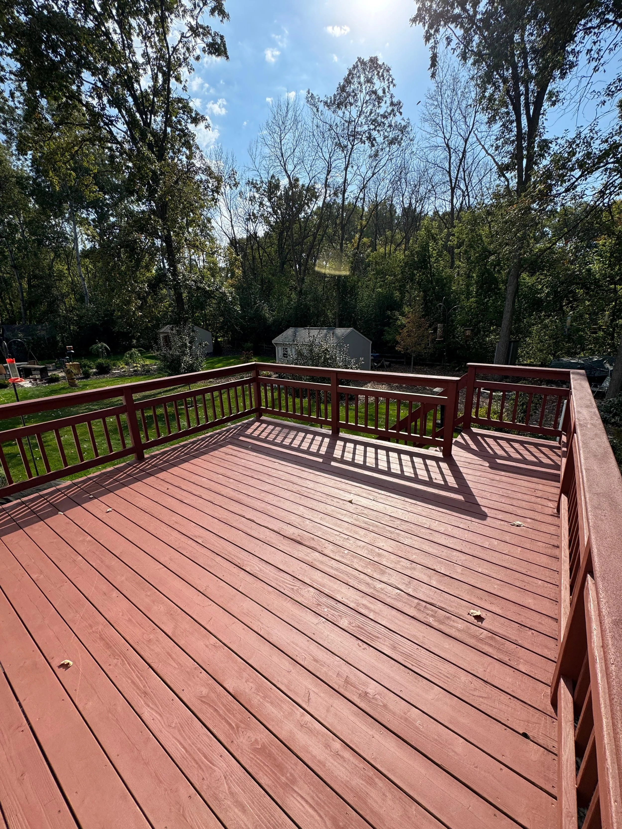 Red wooden deck with railing in a backyard, surrounded by trees, with sheds and outdoor furniture in the background under a partly cloudy sky.