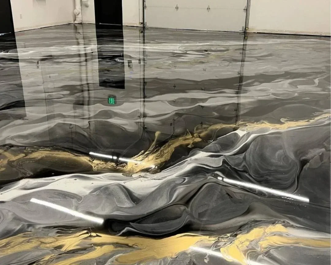 Interior of a building with a reflective, metallic-looking floor that appears to be water or a liquid surface with swirling patterns in black, silver, and gold hues.