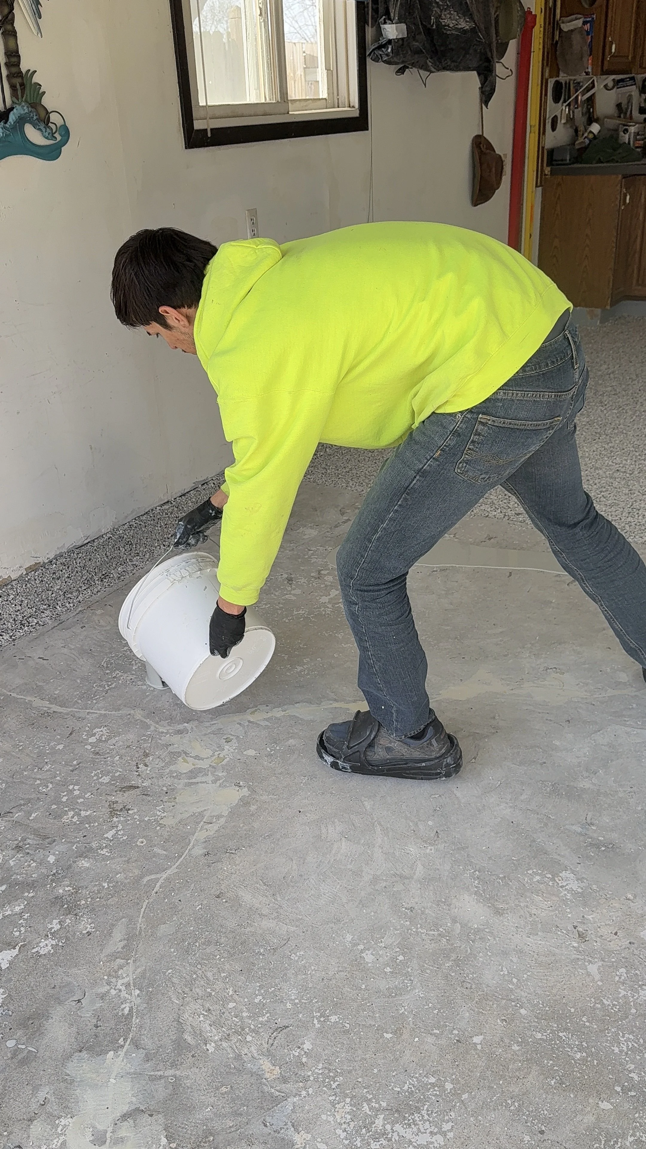 A man wearing a bright yellow hoodie, jeans, and work boots is bending over, pouring a substance from a white bucket onto a concrete floor inside a room. The room has a window and various tools and items on the shelves.
