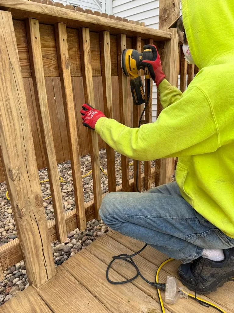 Person wearing a neon yellow hoodie, jeans, and gloves using a power sander on a wooden fence.