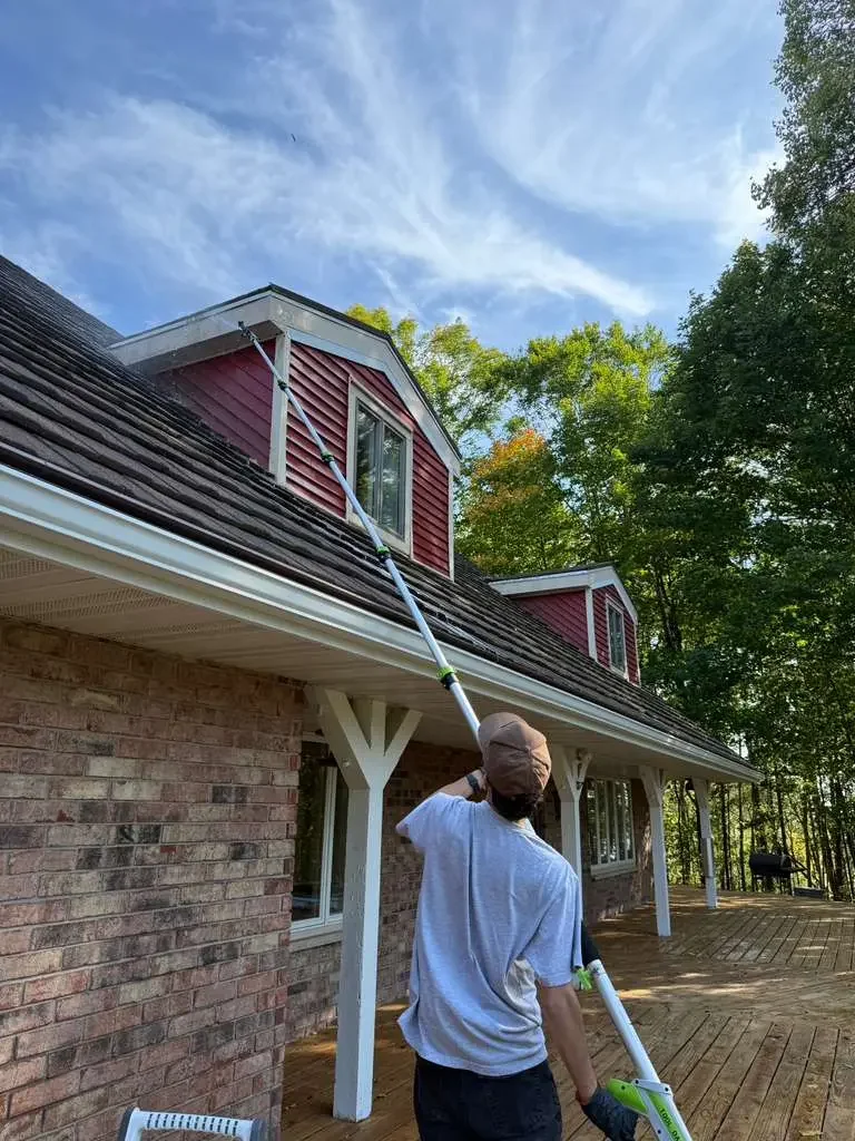 Person cleaning the upper windows of a brick house with a long extendable cleaning tool, on a wooden deck in a backyard surrounded by trees, under a partly cloudy sky.