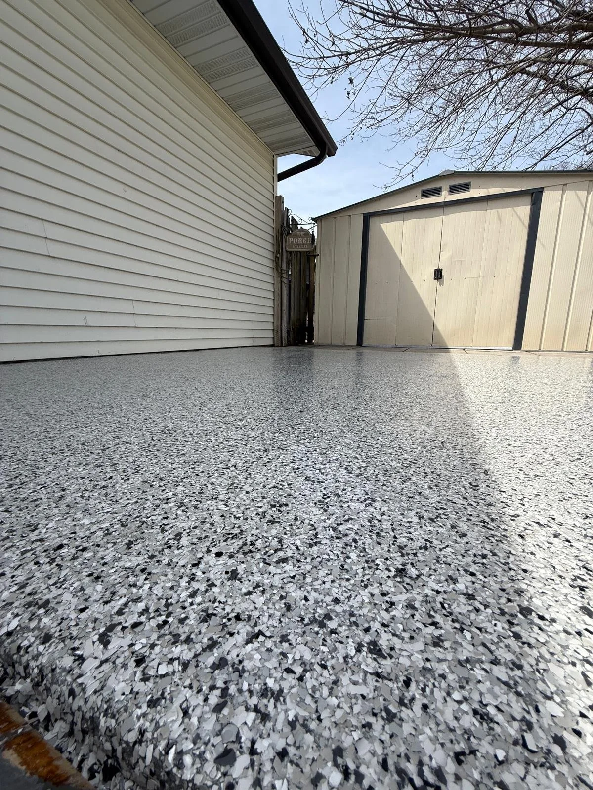 A view of a backyard area with a speckled terrazzo concrete floor, a beige shed with black trim, a wooden fence, and part of a house with cream-colored siding. There is a sign on the fence that says "PORCH." Bare tree branches are visible in the top 