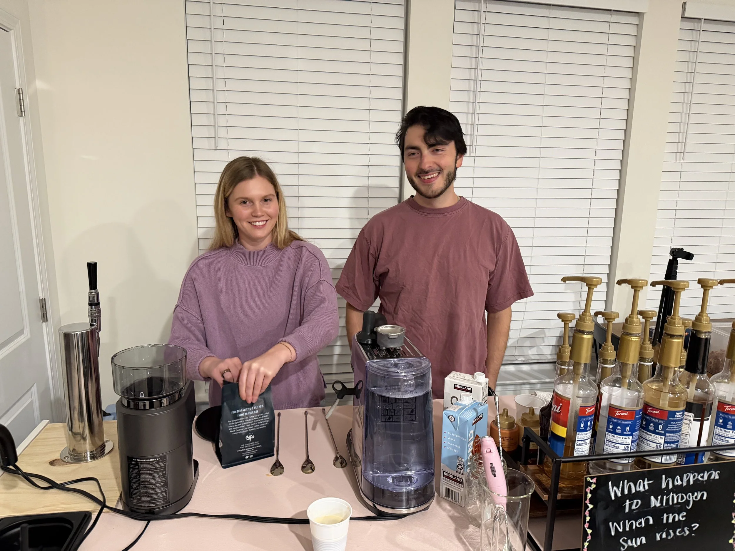 A young woman and man smiling behind a kitchen counter with whipped cream dispensers, syrups, and a sign that reads 'What happens to Nitrogen when the sun rises?'.