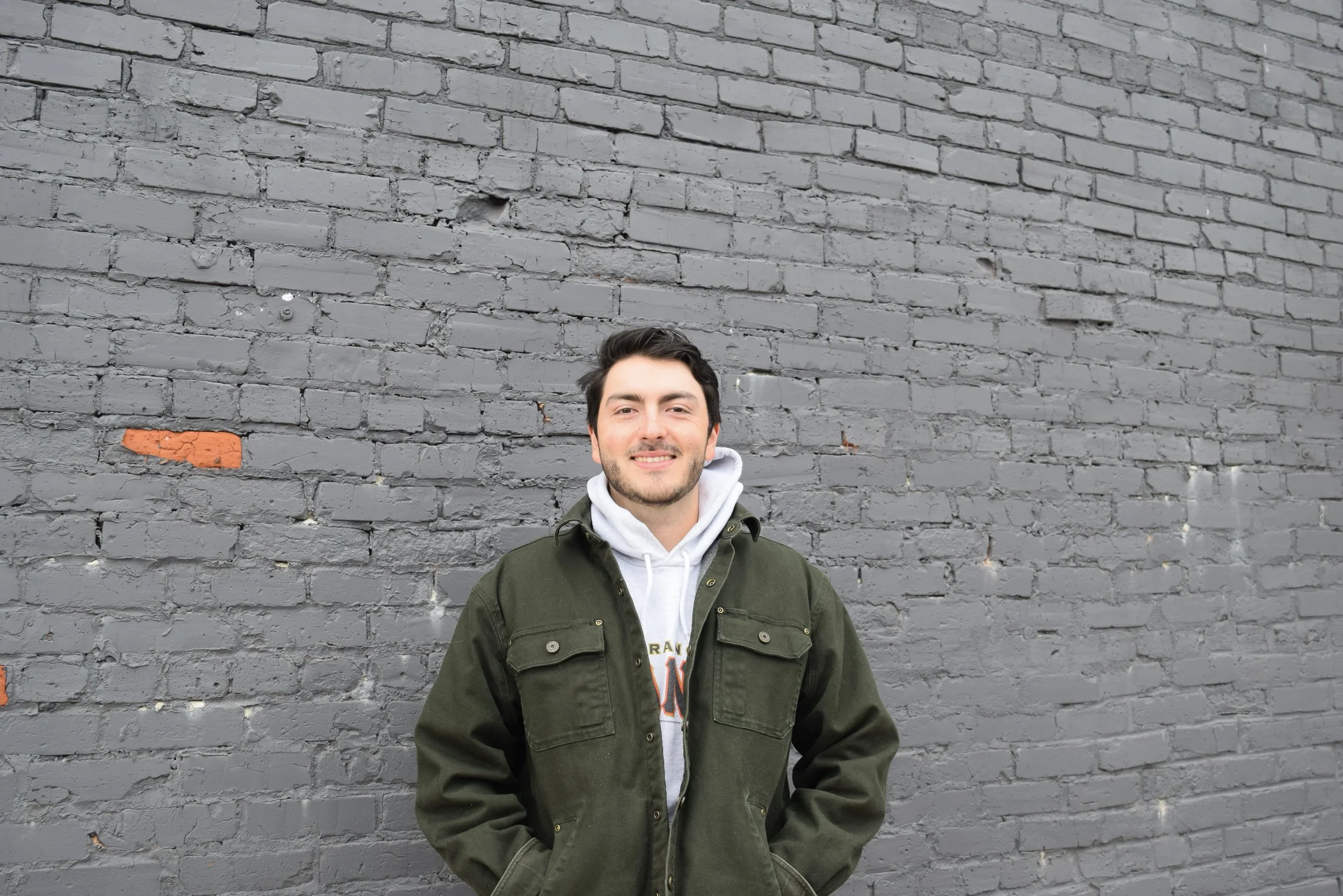 A young man with dark hair and a beard standing in front of a gray brick wall, wearing a green jacket and a white hoodie.