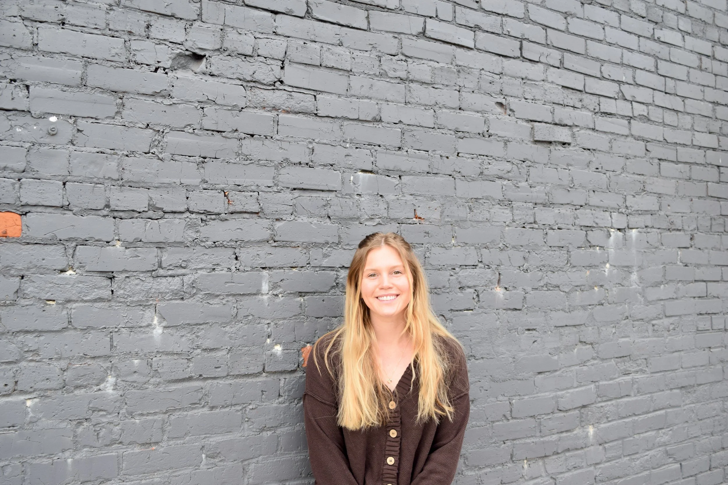 A young woman with long blonde hair and a brown cardigan, smiling and standing in front of a gray brick wall.