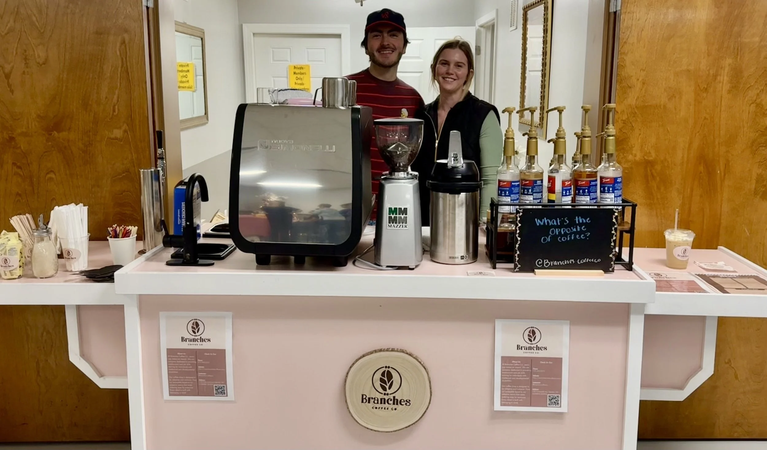 Two baristas standing behind a coffee counter smiling. The counter has coffee equipment, syrup bottles, and utensils. There are signs on the front and a small chalkboard sign asking, 'What's the opposite of coffee?'.