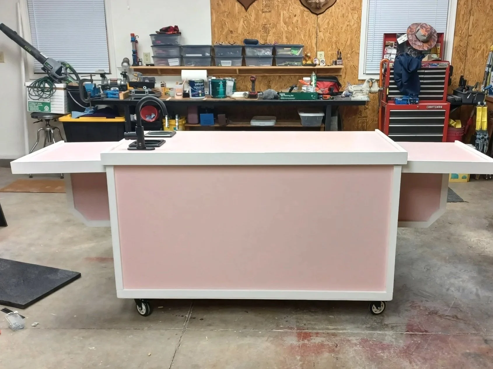 A pink rolling work table with white trim and sides in a workshop, with tools and supplies on a workbench in the background.
