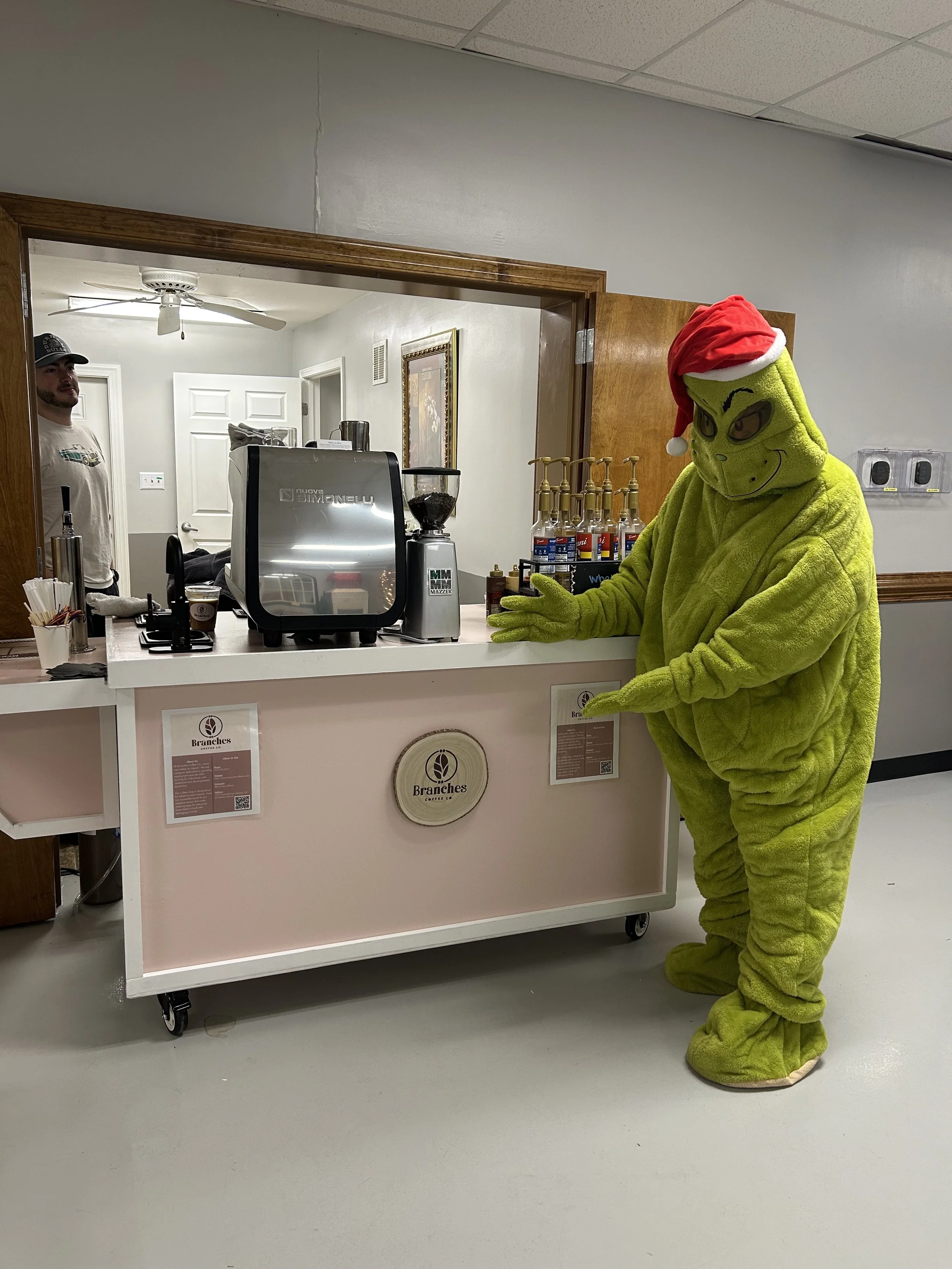 A person in a bright green Grinch costume with a Santa hat standing at a coffee shop counter, handing over an item to a customer.