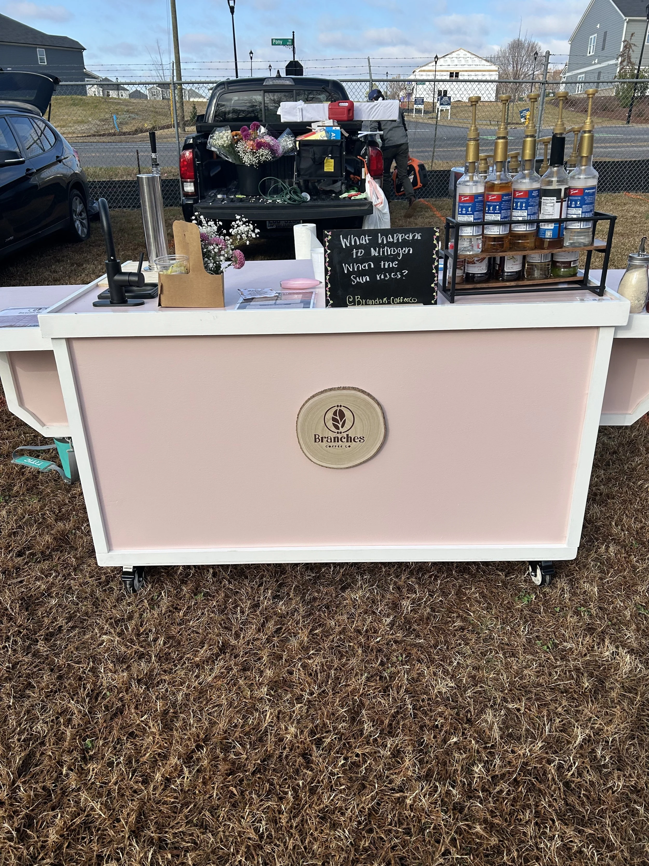 A pink coffee cart with a logo for Branches Coffee Co., set up outdoors on a grassy area. The cart has a floral arrangement, several bottles of alcohol, and a blackboard sign with a question about hydrogen and the sun.