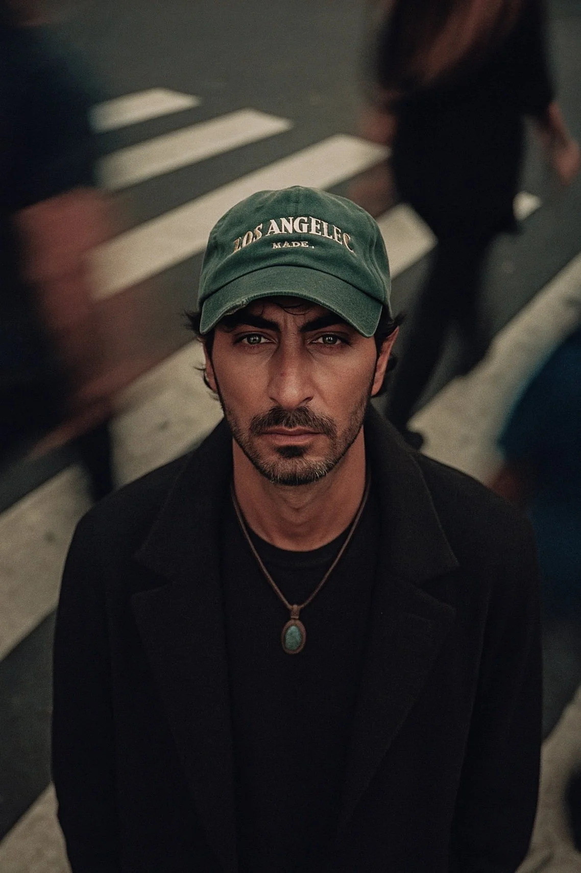 A man with dark hair and a beard, wearing a green baseball cap that says 'LOS ANGELES' and a black coat, stands on a city sidewalk at night.
