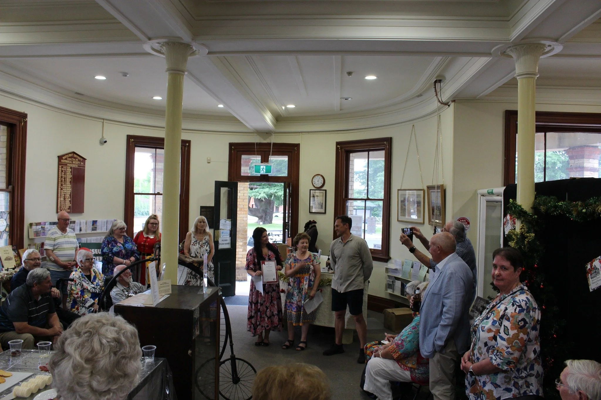 A group of people gathered in a room with wood-framed windows, some standing and some sitting, participating in an event with a speaker and a framed certificate. One man is taking a photo, and there is a decorated Christmas tree on the right.