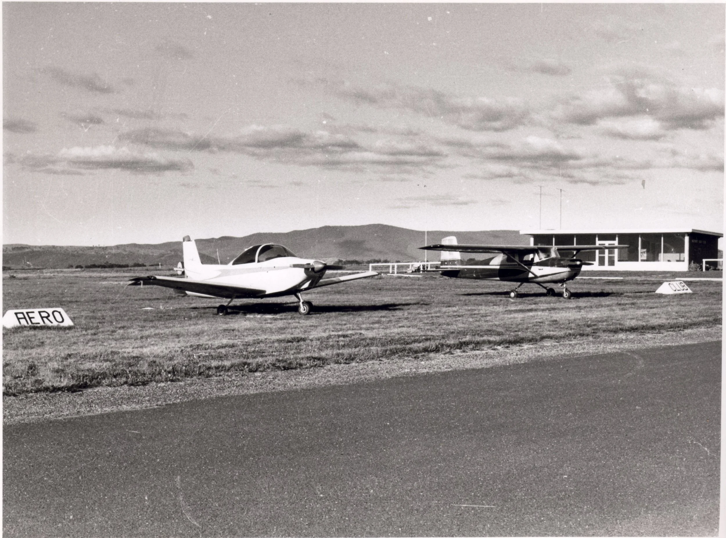 Two small aircraft parked on a grassy airfield with a building in the background and mountains in the distance, under a partly cloudy sky.
