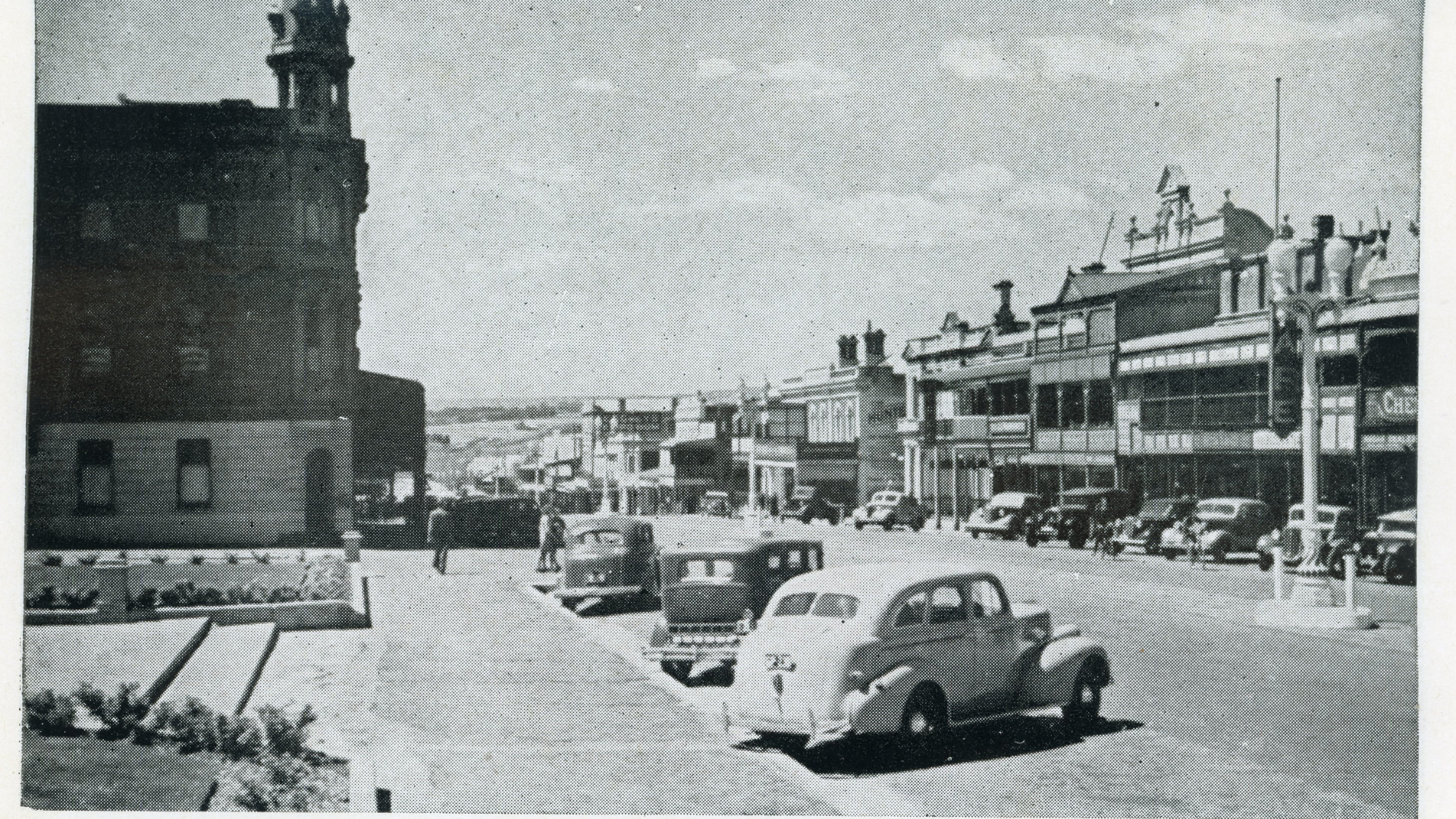 Black and white photo of a vintage street scene with parked cars and businesses with signs on a sunny day.