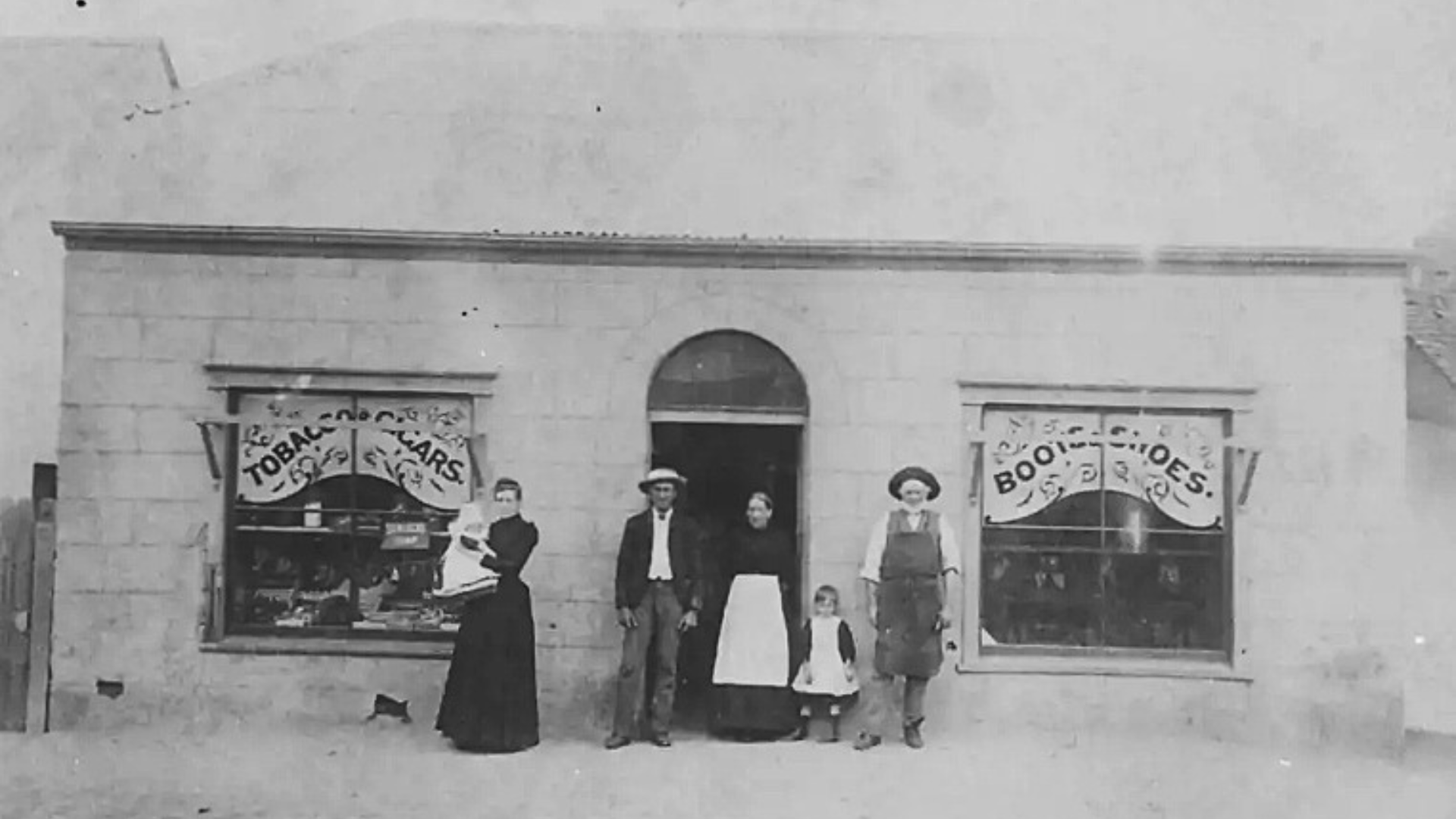 A historic black and white photograph of a small store with three windows and a door in the center. The store sells tobacco, cigars, and bootleg cigarettes. Four people, including a woman with a child, stand in front of the store.