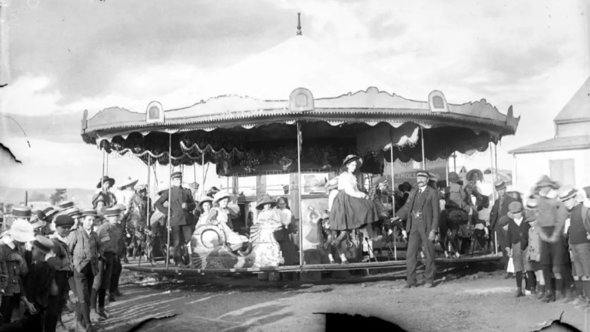 Black and white photo of a vintage carousel with children and adults around it, some riding, some watching, with a large outdoor setting in background.
