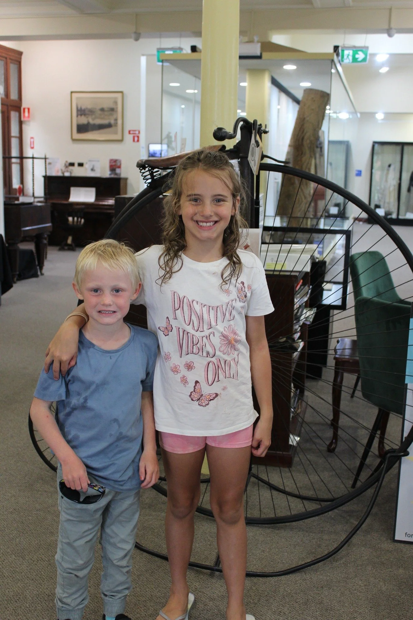 Two children, a boy and a girl, are standing in a museum next to a large vintage bicycle display. The girl has curly hair, is smiling, and is wearing a white t-shirt with pink shorts. The boy has short blond hair, is smiling, and is wearing a blue t-