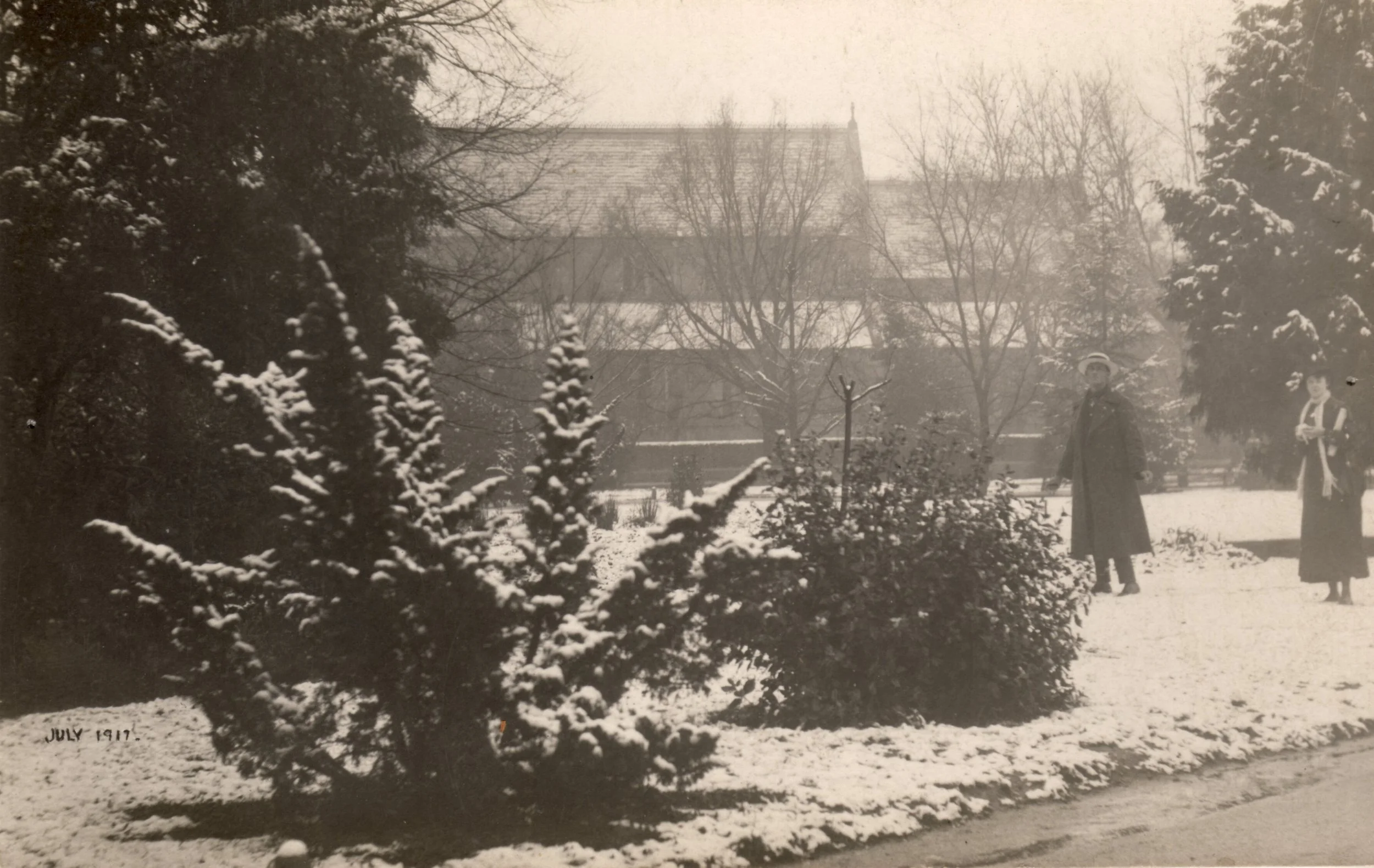 Black and white photograph from July 1917 showing snow-covered bushes and trees in a park, with two women dressed in long coats and hats standing and walking along a snow-covered path, with a large building in the background.
