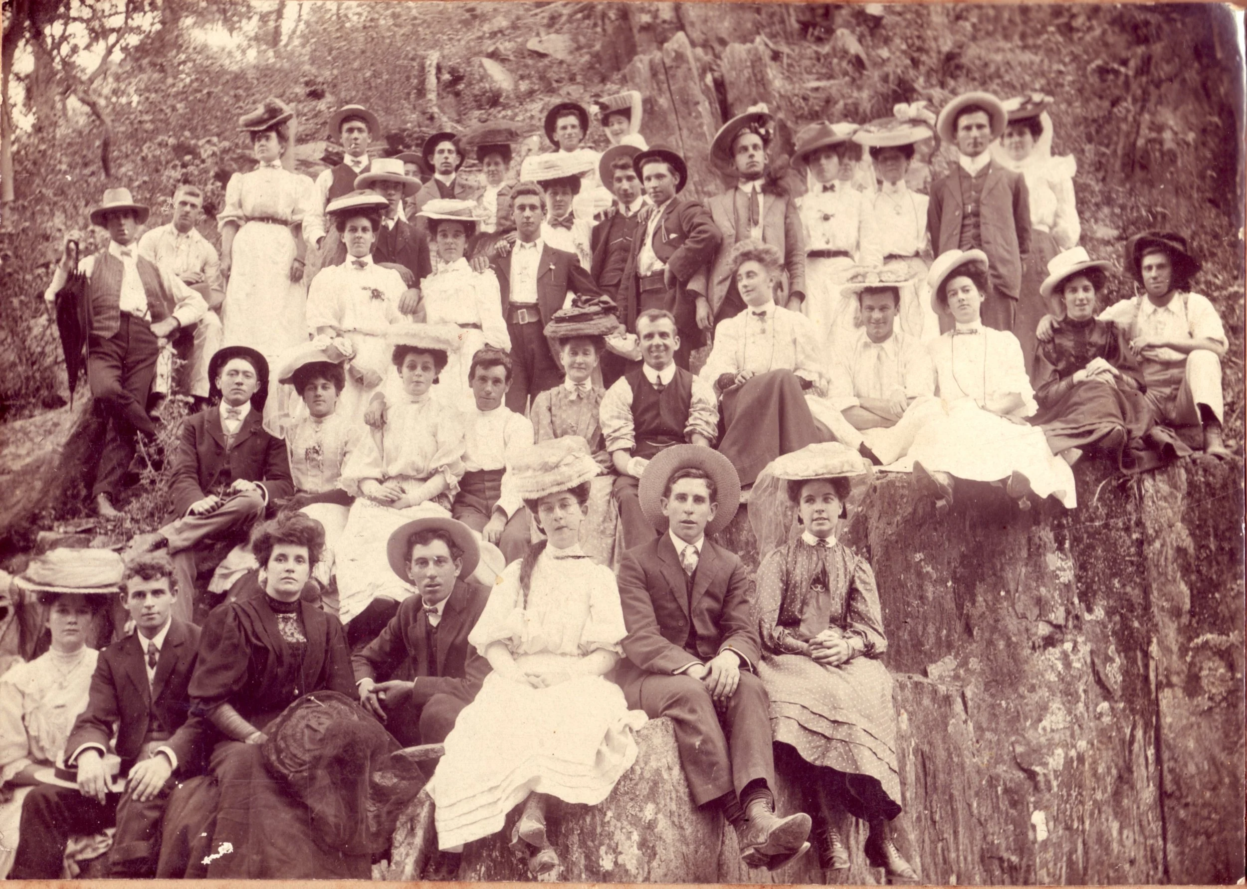 A vintage black-and-white photograph of a large group of men, women, and children posing outdoors on a rocky hillside, dressed in early 20th-century clothing with women in long dresses and wide-brimmed hats, and men in suits and hats.