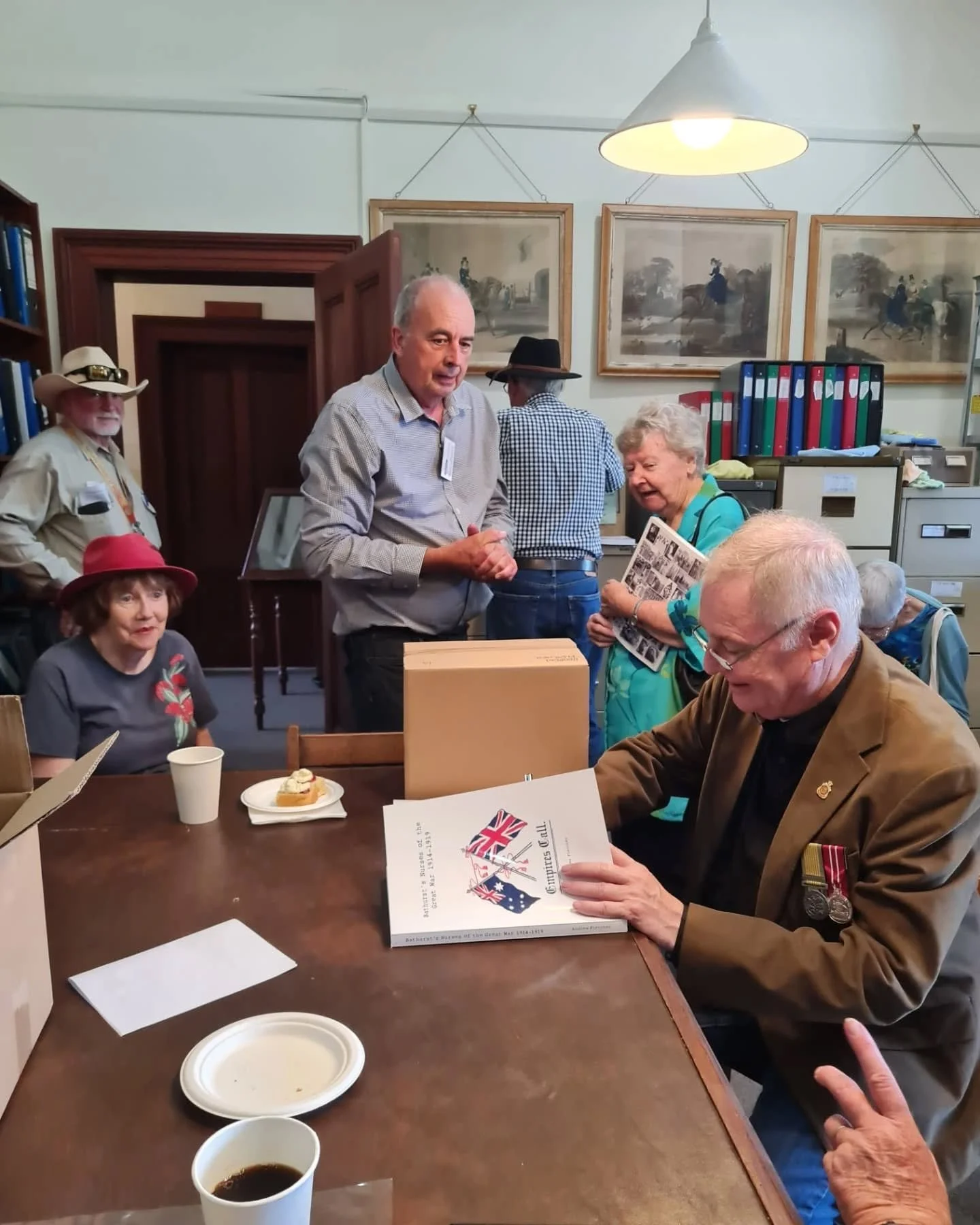 Group of elderly people gathered in a room, some seated at a table. One man is sitting and looking at a book, an older woman is standing and laughing, and others are standing and talking. The room has framed paintings on the wall, a hanging light, an