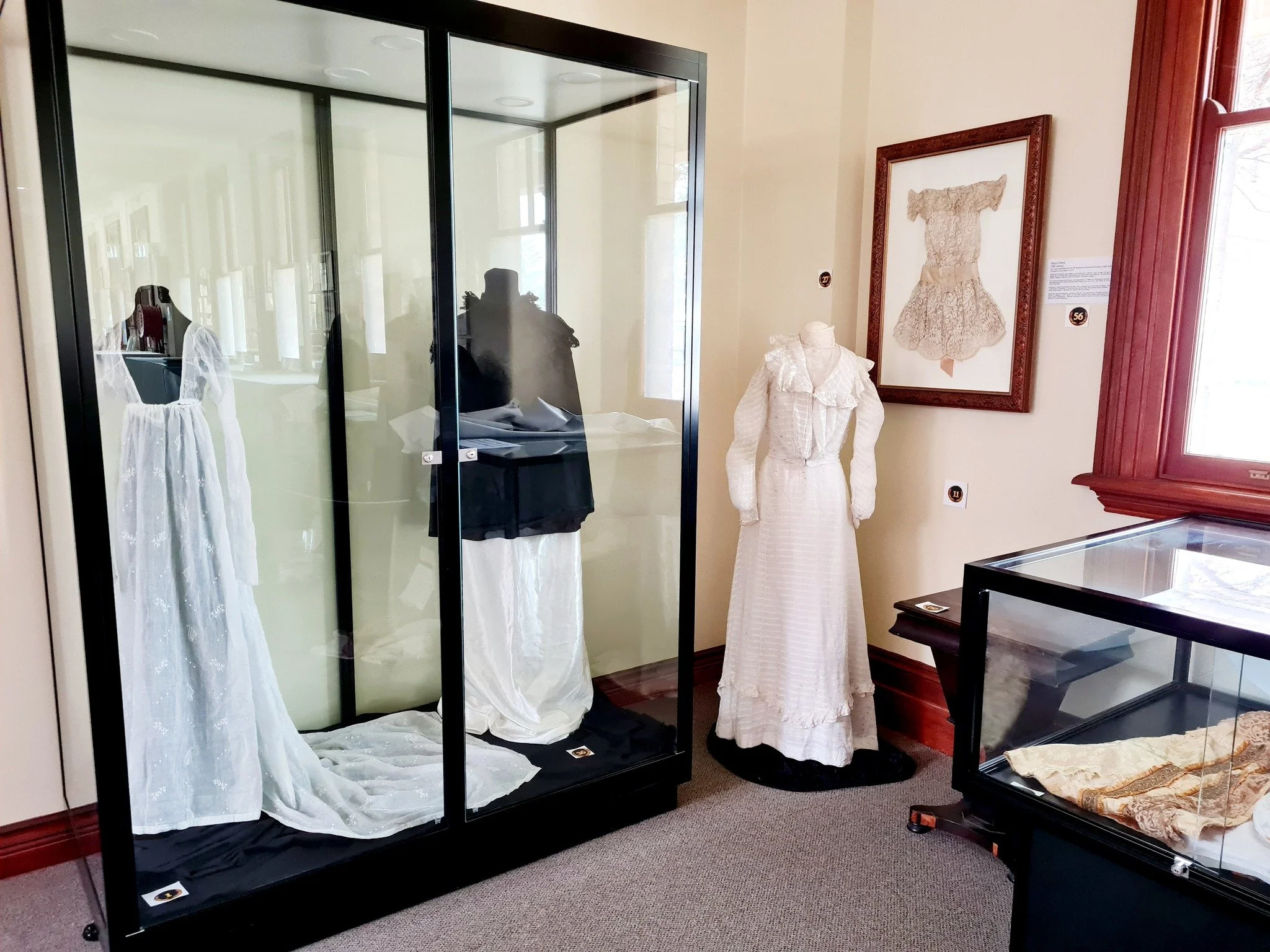 Museum display of vintage dresses in glass cases, with one dress on a mannequin and framed artwork of a lace dress on the wall.