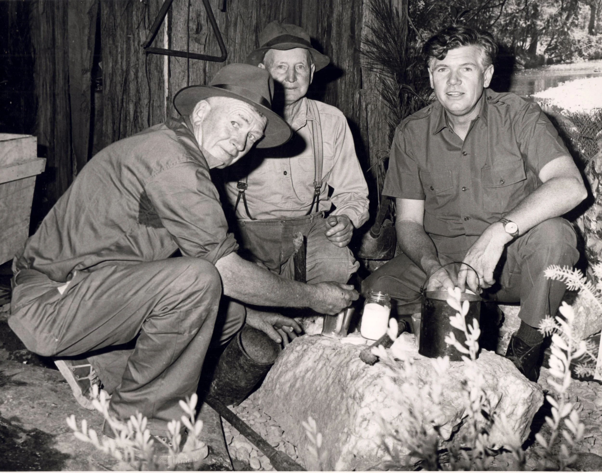 Four men in outdoor clothing and hats around a campfire, with a wooded background. They are sitting and tending to the fire, with jars and a small axe visible.