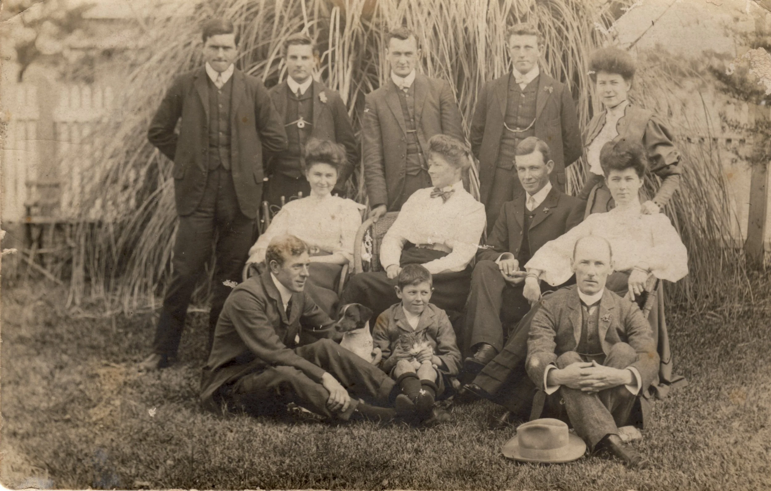 A vintage black-and-white photograph of a large family outdoors, with several adults and children arranged in front of tall plants or grass. Some individuals are seated, others standing, and a dog and a cat are also present.