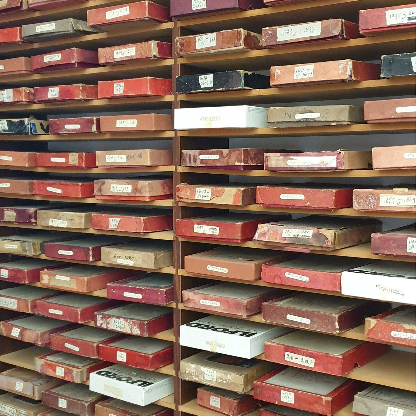 Multiple shelves filled with boxes, many are red, some are white and beige, labeled with handwritten notes and collection dates. The boxes appear old and stored for archival purposes.