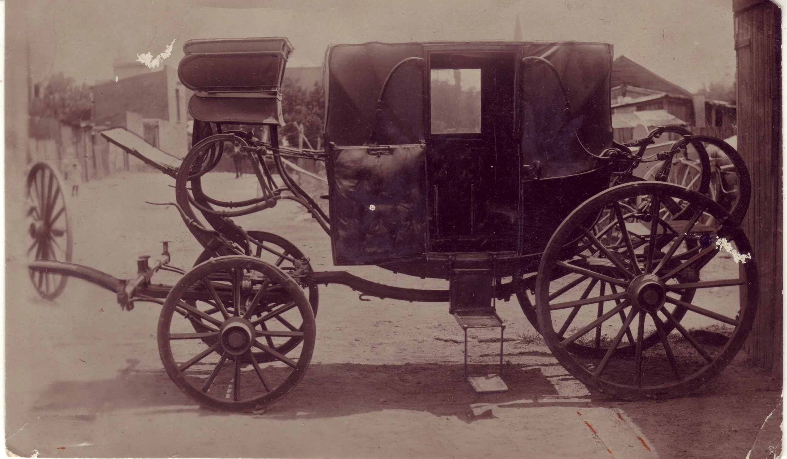 An old black-and-white photo of a horse-drawn carriage with four large spoked wheels, parked on a dirt street with buildings in the background.