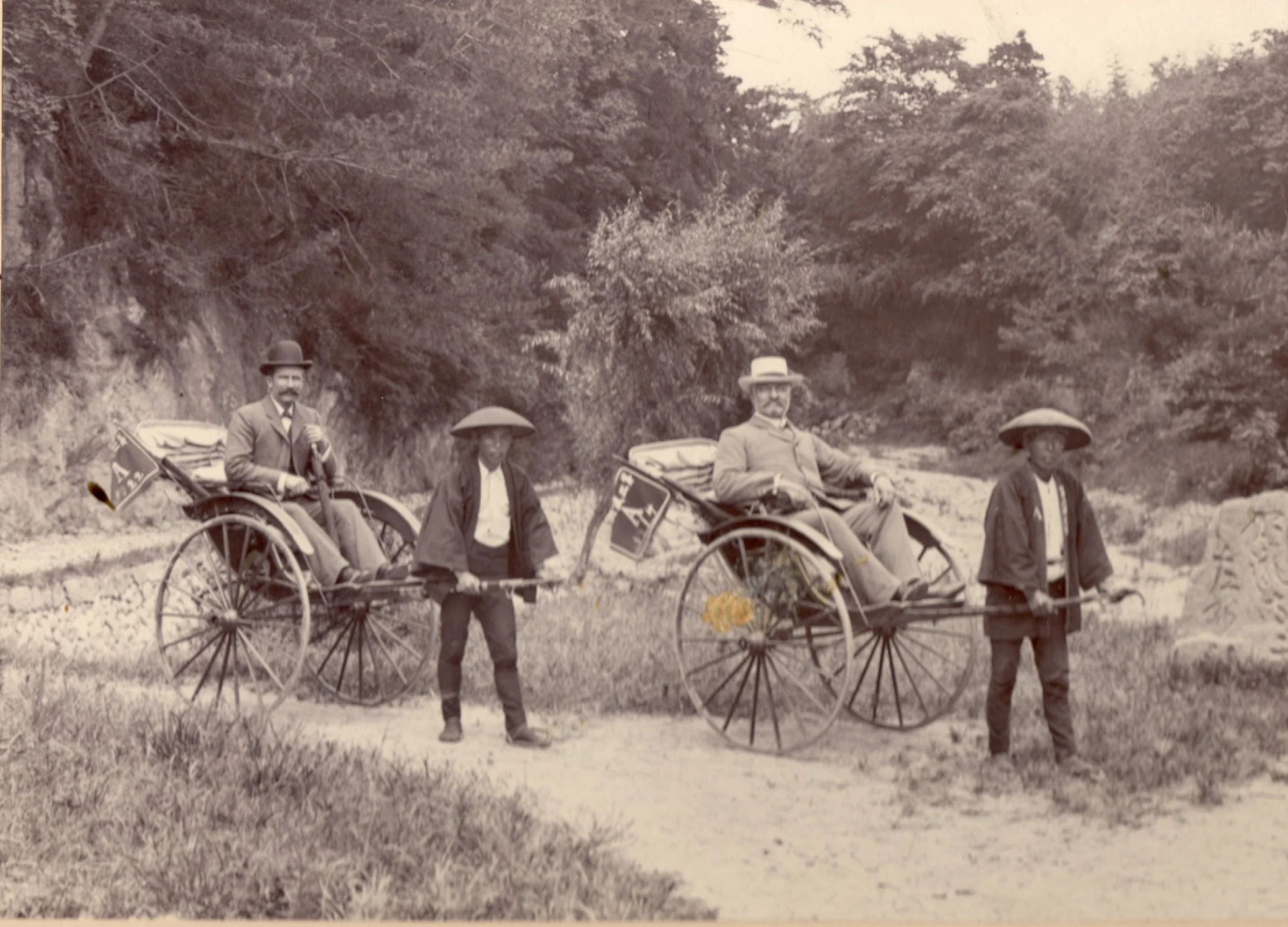Black and white photograph of two men in rickshaws with two men walking beside them, set outdoors with trees and a dirt path