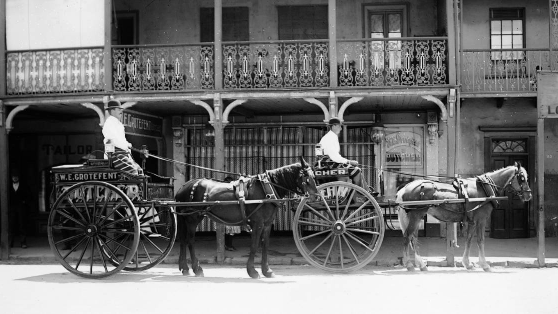 Two horse-drawn carriages, each with a driver, parked in front of a building with balcony and storefronts, in black and white.