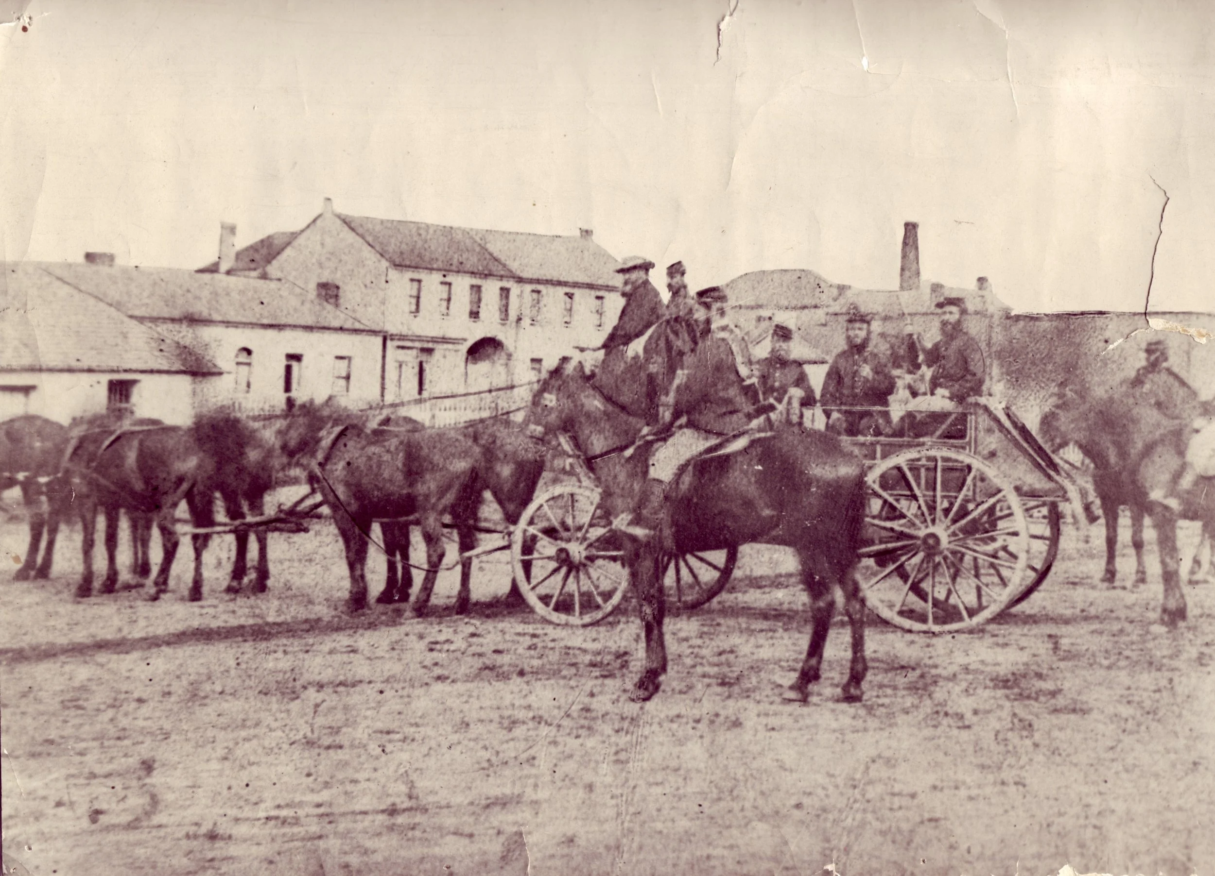 An old sepia photograph showing a group of men riding in a horse-drawn wagon and standing around it, with several horses hitched to the wagon, in a town square with historic buildings in the background.