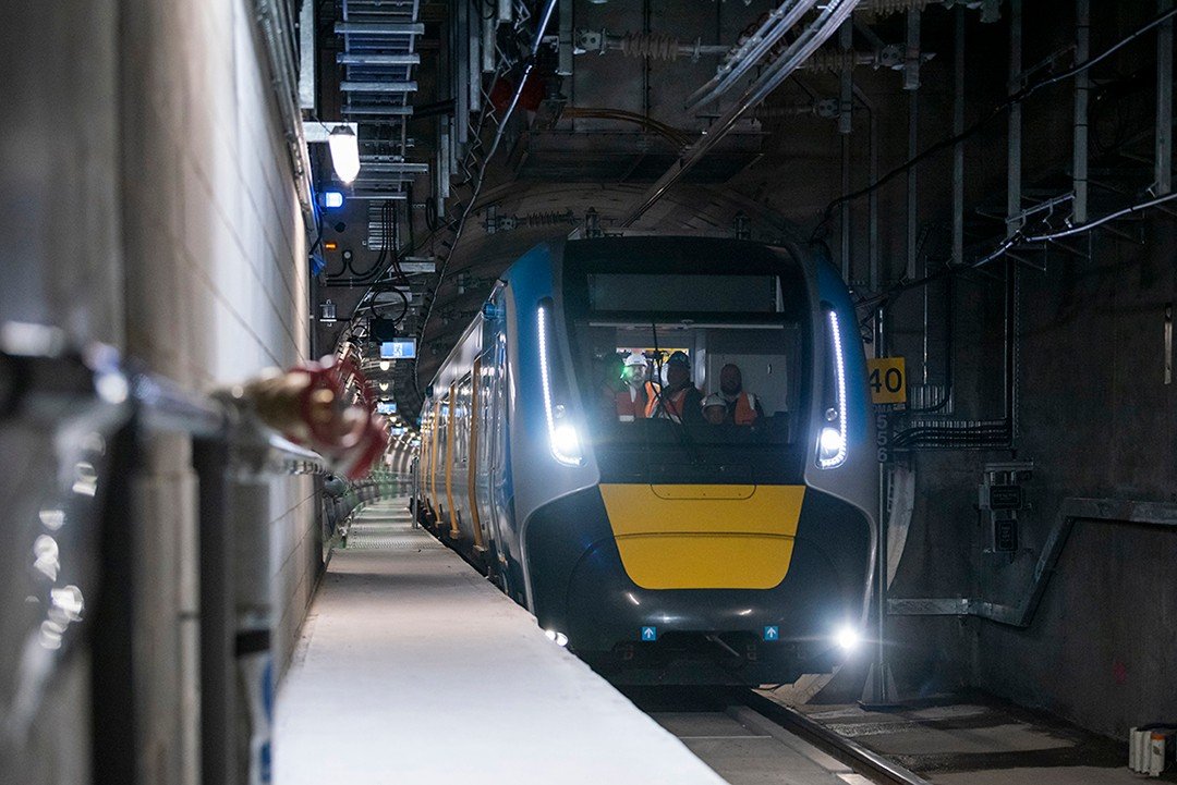 Modern train with bright headlights entering underground tunnel, construction workers on board, illuminated equipment and cables overhead.