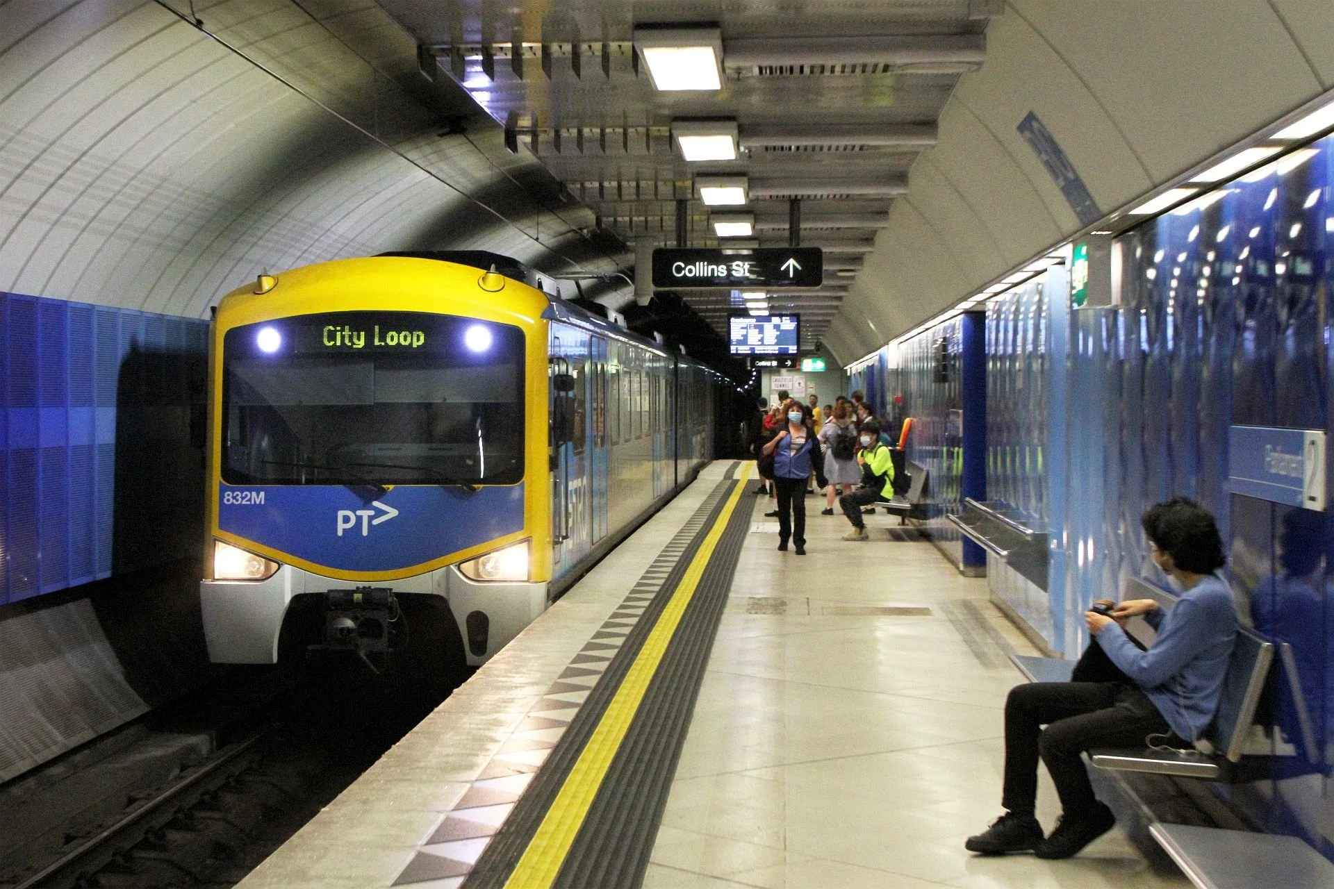 People waiting on a subway platform for a city metro train labeled 'City Loop'. The train is approaching the platform, and some passengers are seated while others are standing or walking. The station sign shows 'Collins St'.