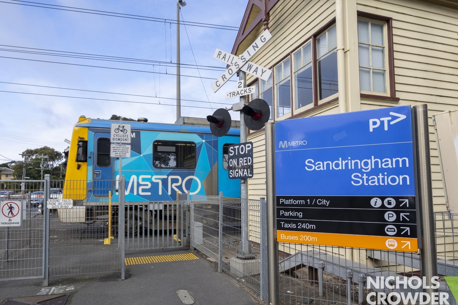 Sydney Metro train at Sandringham Station with station sign, railway crossing signals, and surrounding fencing and signage.