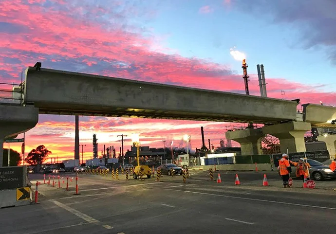 Construction workers and vehicles near an overpass at sunset, with pink and blue sky above.