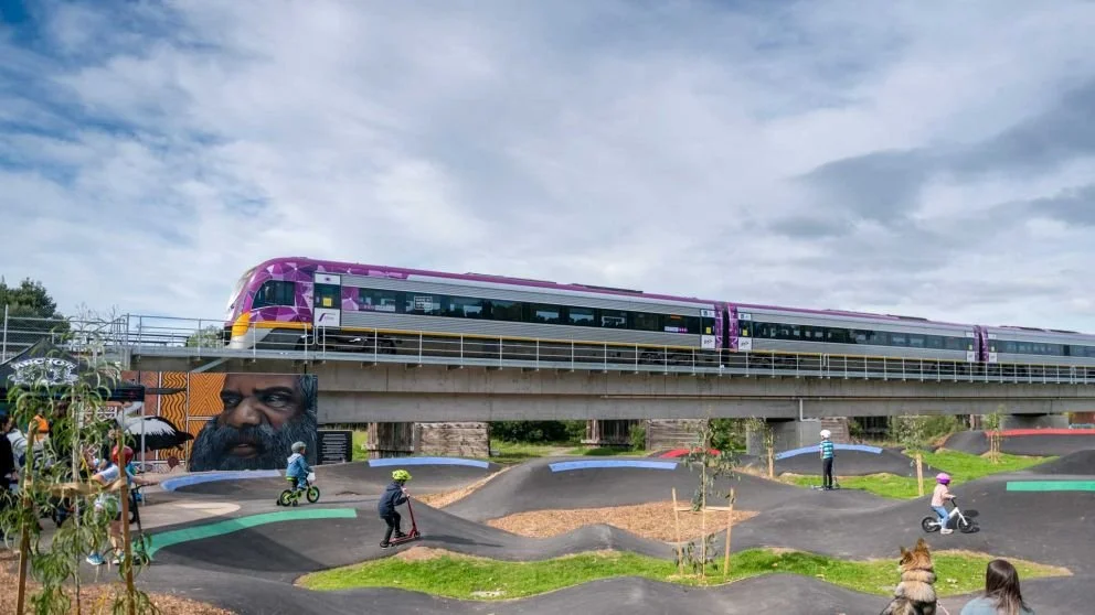A light rail train traveling on an elevated track over a pump track with children riding bikes and scooters underneath, and murals on the walls.