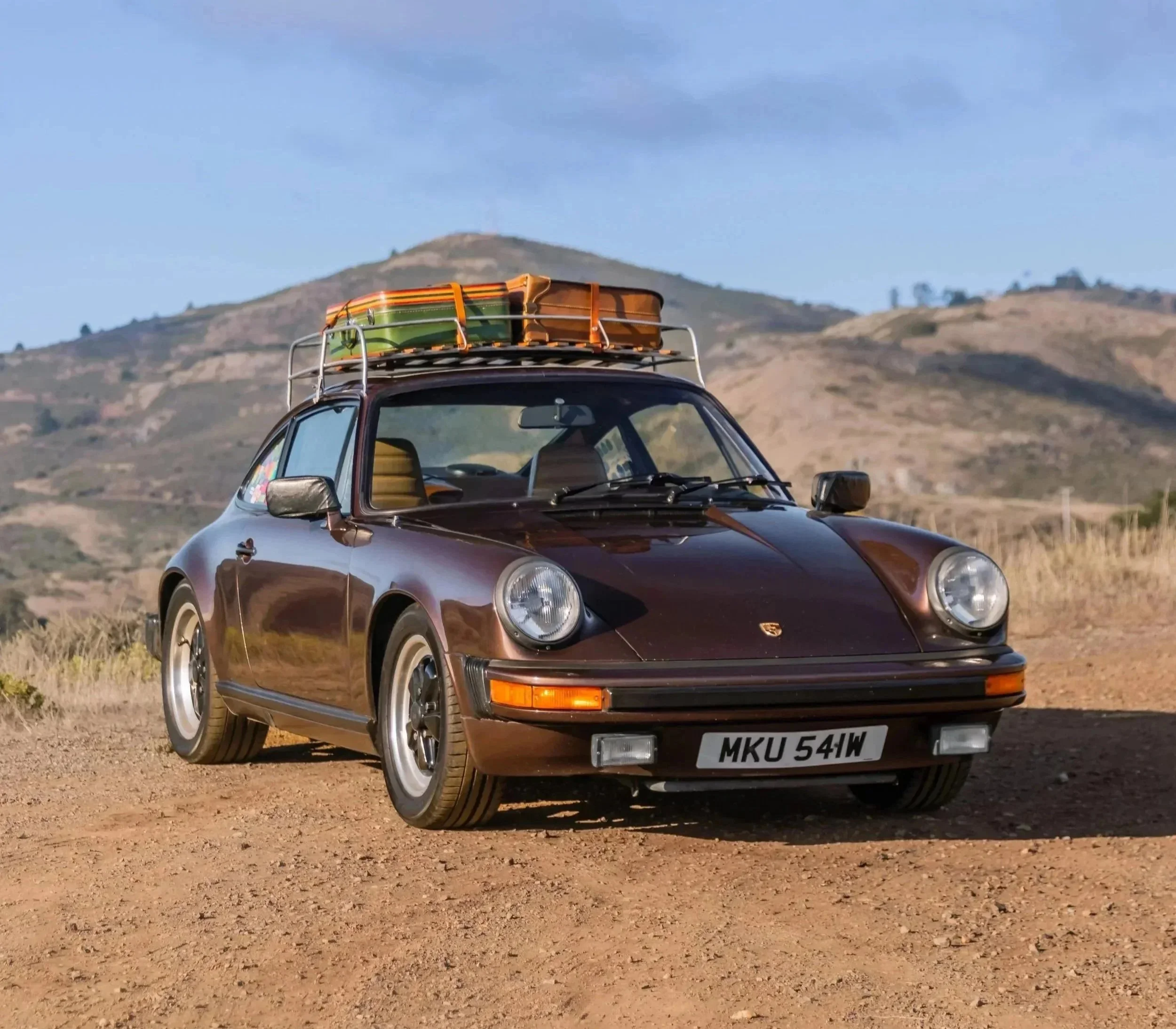A vintage brown Porsche 911 car parked on dirt ground with luggage racks on the roof carrying colorful suitcases, hills and a blue sky in the background.