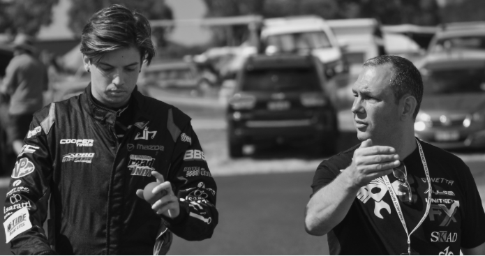 Two men in a parking lot, one looking at a phone and the other gesturing with his hand, with cars in the background.