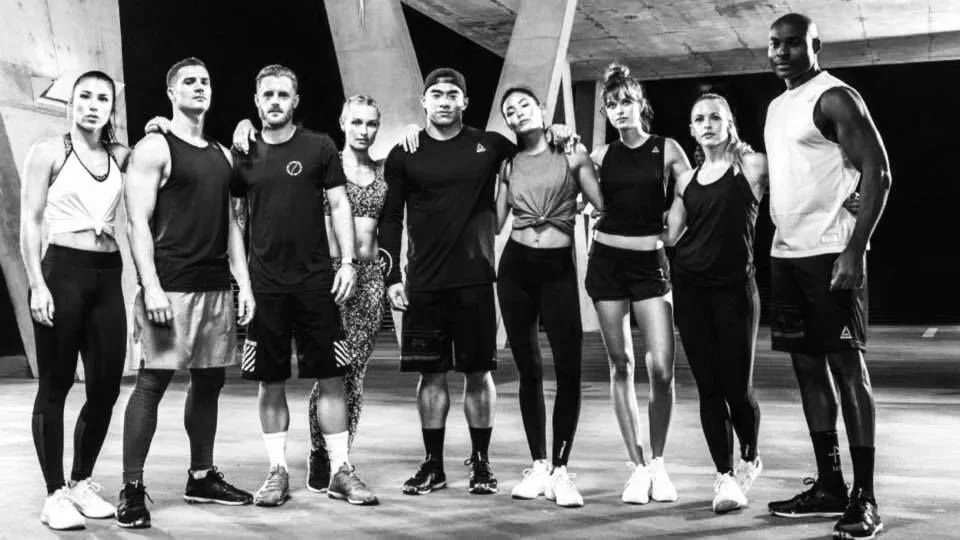 A group of nine diverse young adults standing together in an indoor gym or fitness studio with modern architecture, dressed in athletic clothing, posing for a photo.