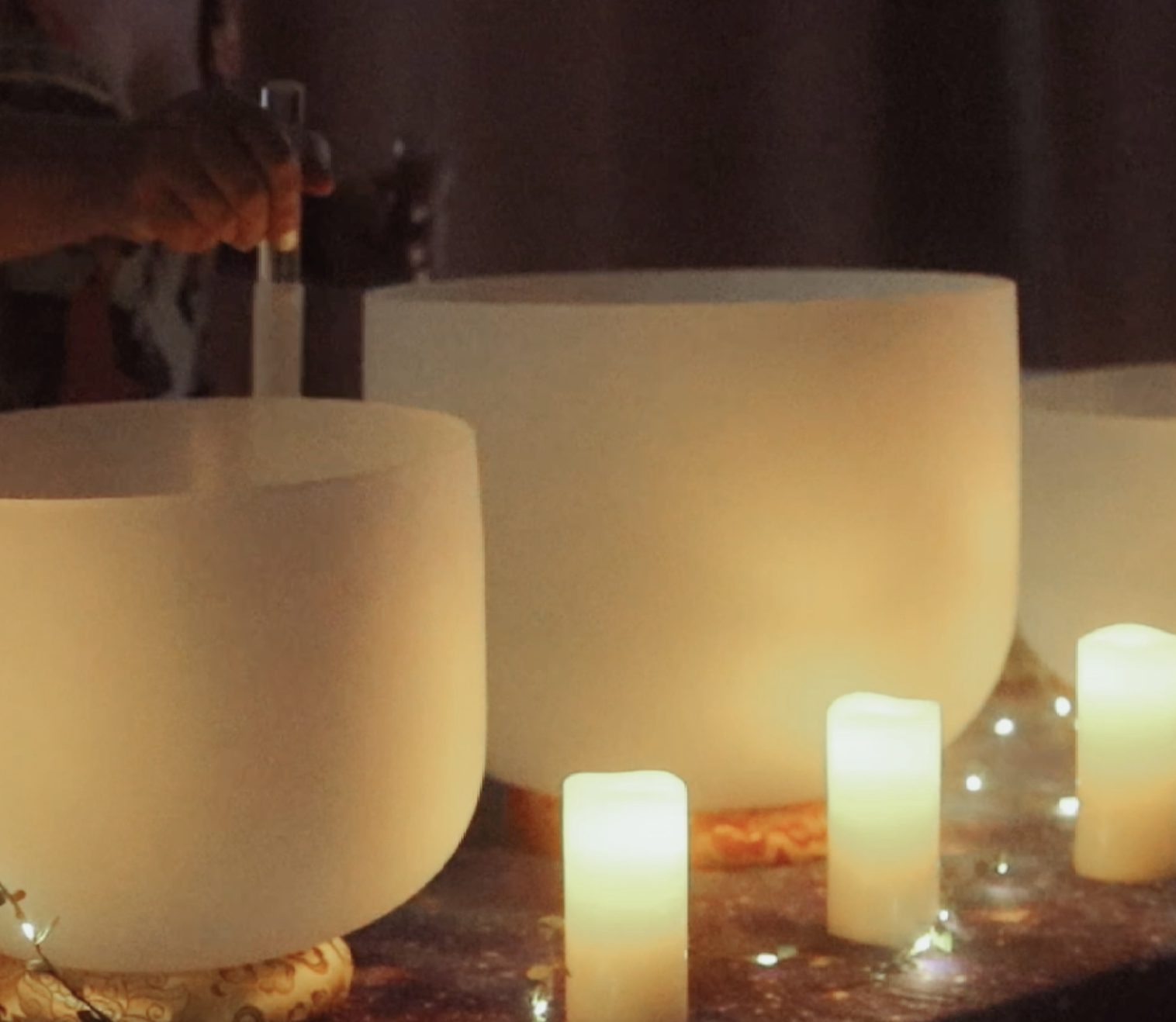 A person playing a singing bowl with a wooden mallet surrounded by lit candles and fairy lights in a cozy setting.