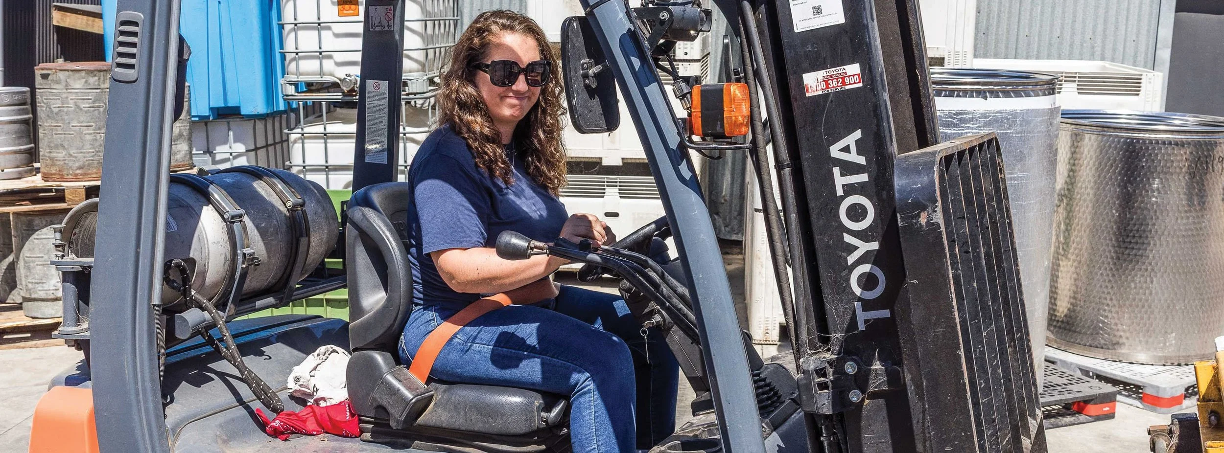 A woman with curly hair and sunglasses operating a Toyota forklift in an outdoor warehouse or industrial area with metal containers and equipment in the background.
