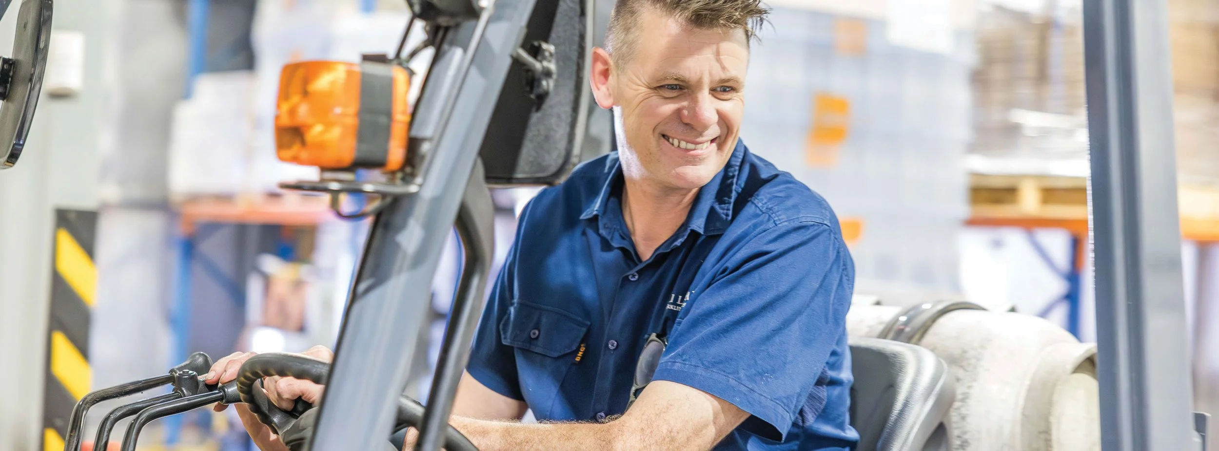 A smiling man in a blue uniform operating a forklift in a warehouse.